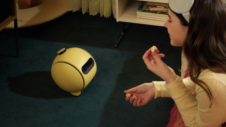 Samsung's yellow ball-shaped Ballie robot rolls across a dark green carpet toward a girl kneeling with popcorn in her hands.