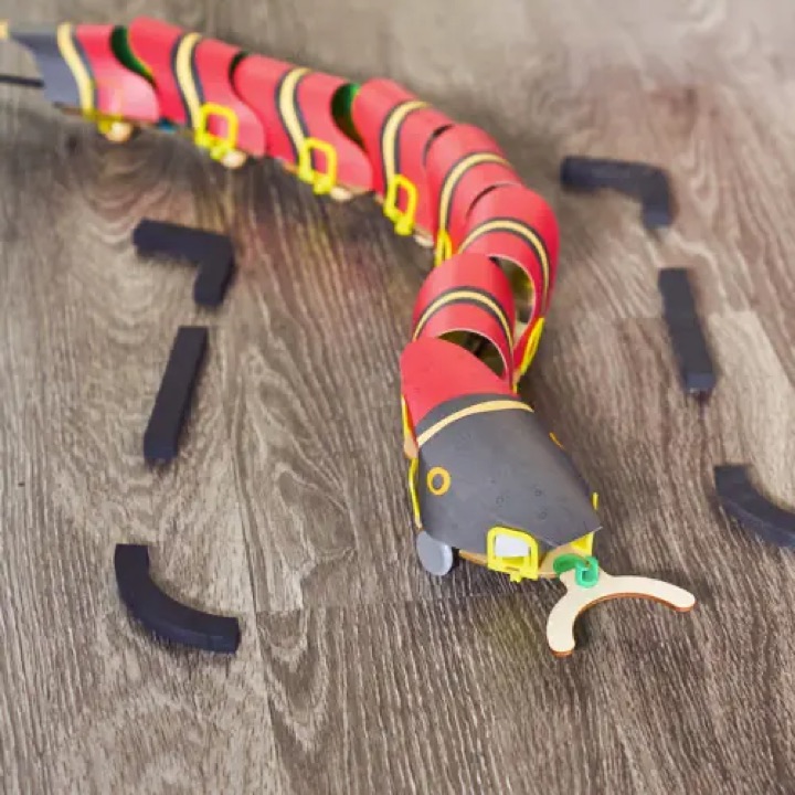 Assembled KiwiCo remote-controlled snake robot with red segmented skin, a dark head, and a wooden wishbone-shaped tongue slithering across a wooden floor between foam obstacles.