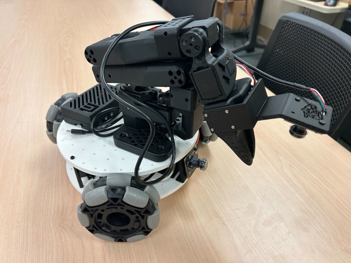 Assembled LeKiwi robot on a wooden desk: a white circular base on three omni wheels, topped by a black 3D-printed arm with a two-finger gripper and a small front camera board.