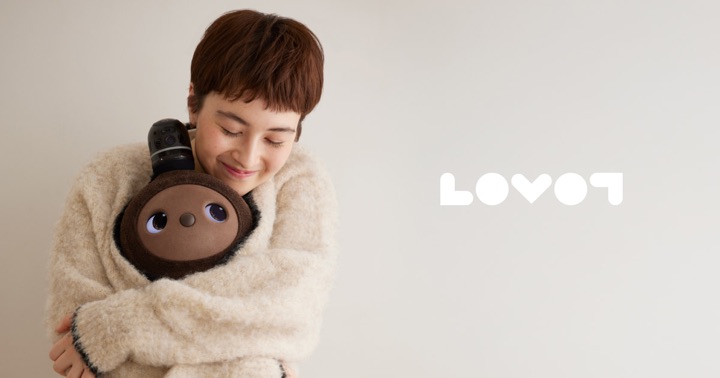 A short-haired woman in a cream sweater hugs a brown LOVOT robot with large dark eyes against a beige wall, with the LOVOT wordmark at right.