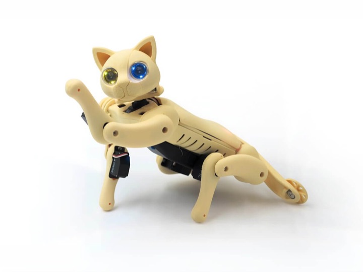 The cream-colored Petoi Nybble Q robot cat with one yellow and one blue LED eye kneels on its front legs and raises a paw on a white background.