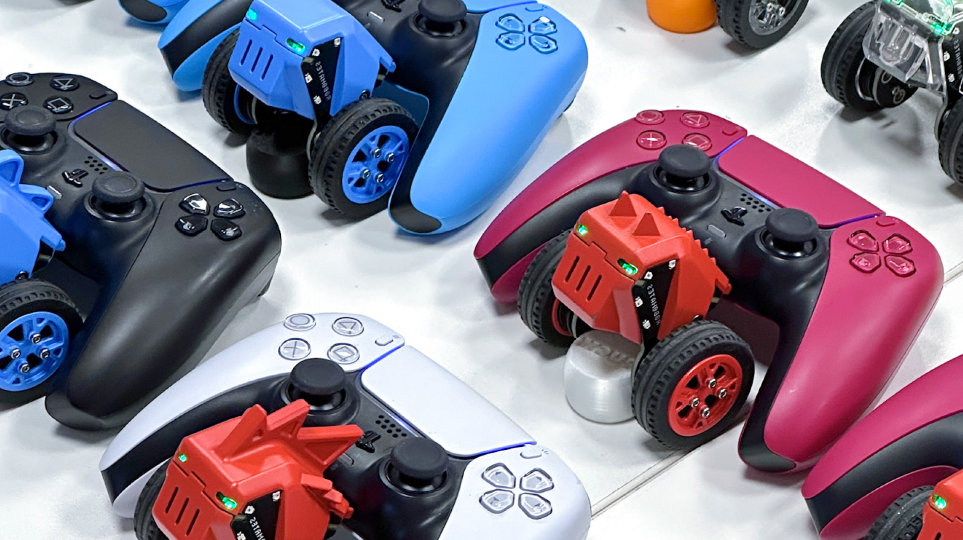 Lifestyle shot: several assembled Robomates in blue and red parked beside PS5 DualSense controllers on a white table, status LEDs on.