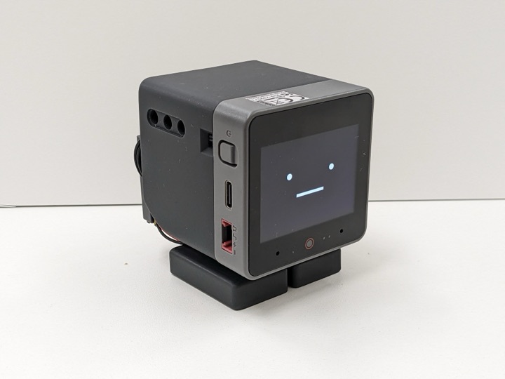 Assembled Stack-chan robot, a palm-sized cube built around an M5Stack module showing a minimal face on its screen, with dark gray shell and small black feet on a white table.